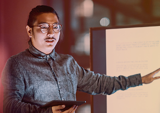 A person in a grey button-up shirt with glasses gestures towards a presentation screen while holding a tablet.