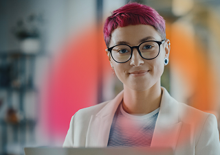 A person with short pink hair and glasses smiles while looking at a laptop, wearing a white blazer over a striped shirt.
