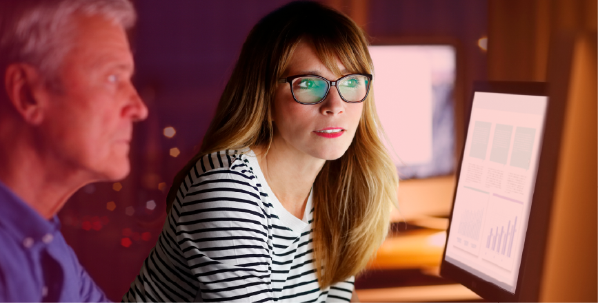 A woman with long hair in glasses discusses data with a man, focusing on a computer screen displaying graphs and charts.