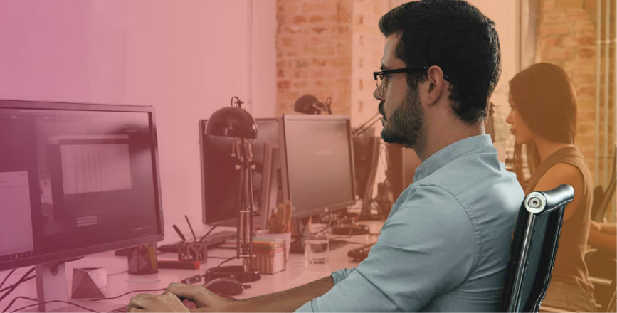 A man with glasses is seated at a desk, focusing on a computer screen, while a woman works in the background.