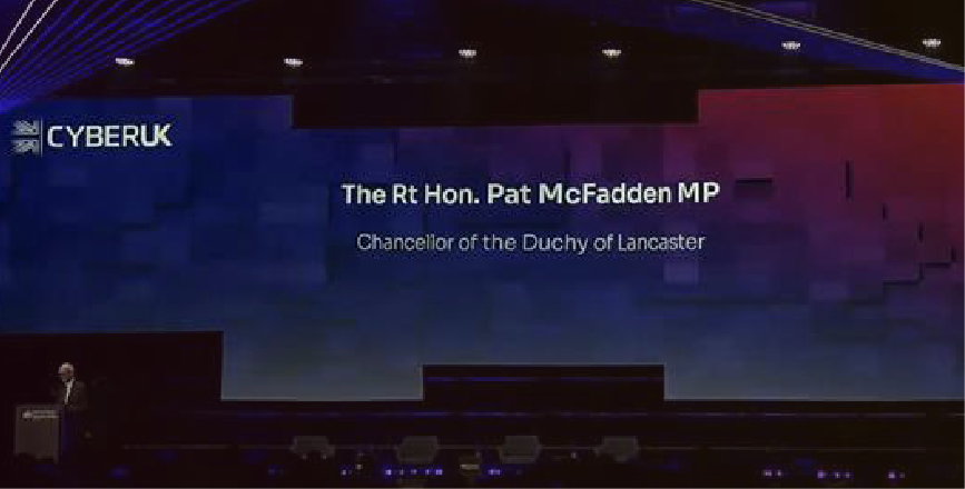A speaker at a conference stands in front of a large screen displaying the name and title of Pat McFadden MP, Chancellor of the Duchy of Lancaster.