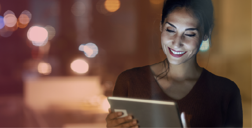 A woman smiling while using a tablet, illuminated by soft lights in a cozy indoor setting.
