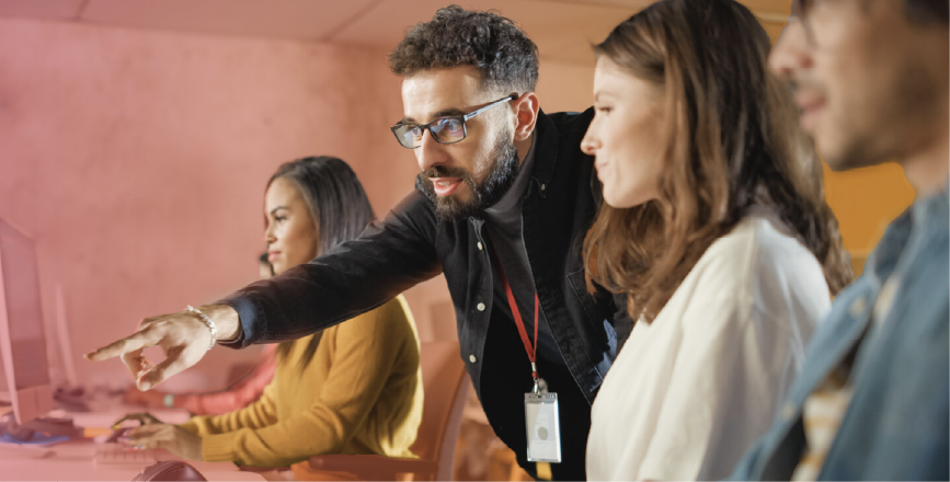 A group of four people working at computers, with one person pointing and explaining something to two others, while a fourth watches.