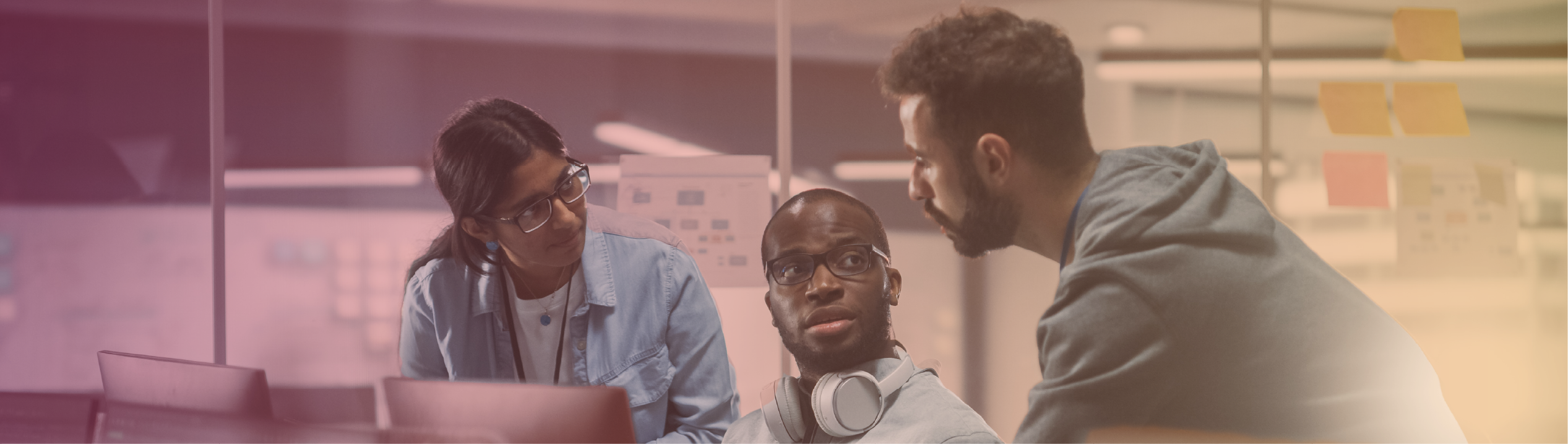 A diverse group of three professionals collaborating in an office, with one person wearing headphones looking attentively at the others.