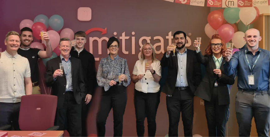 A group of eight people celebrating with drinks in an office decorated with balloons and a logo for 