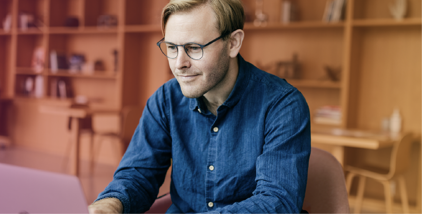 A man with glasses sits at a laptop in a bright, modern workspace, focused on his screen.