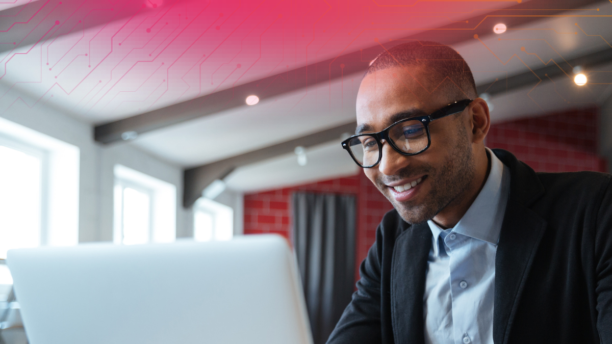A smiling man wearing glasses works on a laptop in a bright, modern office with decorative lighting.
