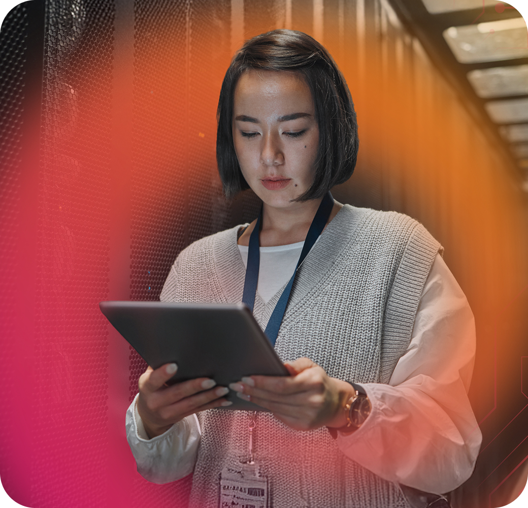 A woman with short dark hair is using a tablet while standing in a server room, wearing a grey sweater and an identification badge.