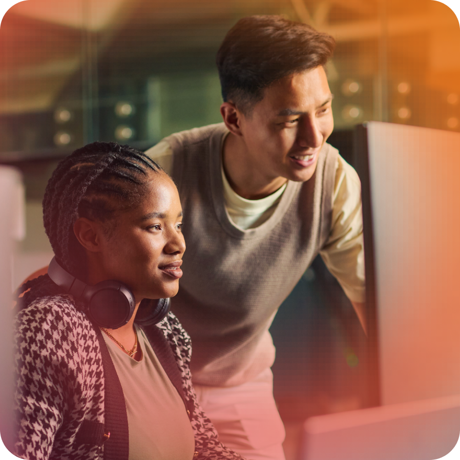 A young woman wearing headphones looks at a computer screen, while a young man stands beside her, smiling and engaged.